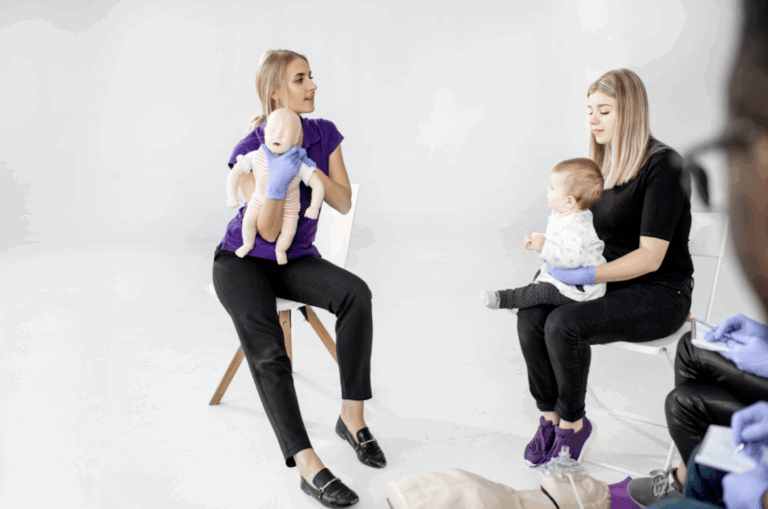 Instruktorė demonstruoja kūdikio užspringimo veiksmus su manekenu, šalia mama su kūdikiu mokymuose.