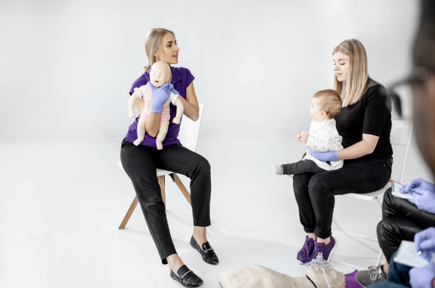 Instruktorė demonstruoja kūdikio užspringimo veiksmus su manekenu, šalia mama su kūdikiu mokymuose.